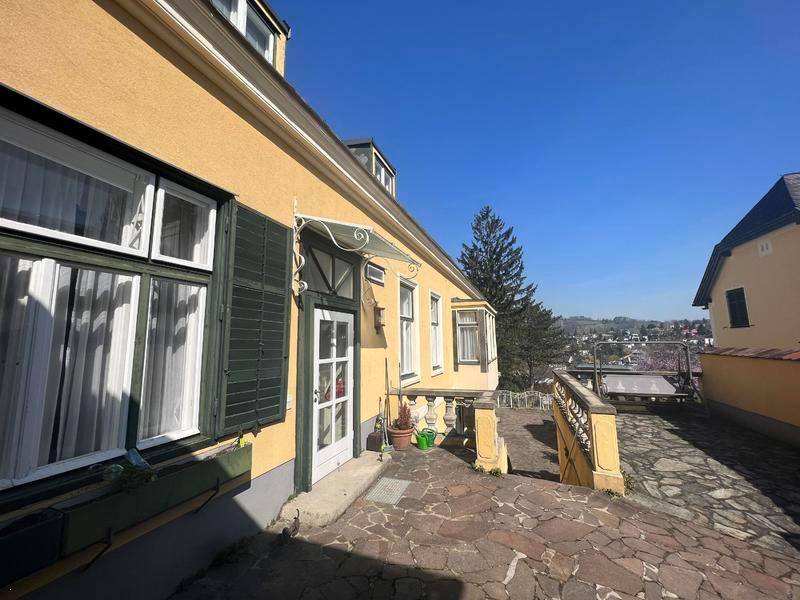 Gelbes Gebäude mit grünen Fensterläden und gepflastertem Eingangsbereich unter blauem Himmel.