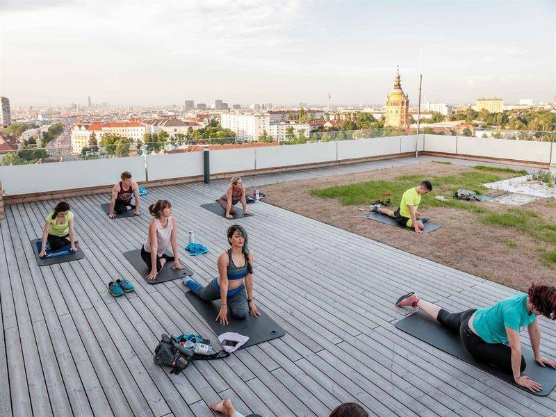 Menschen praktizieren Yoga auf einer weitläufigen Dachterrasse mit Holzboden und Panoramablick über die Stadt.