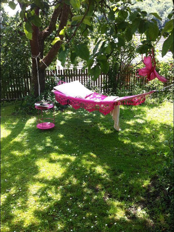 Sonnige Hängematte mit rosa Fransen unter einem Baum im grünen, blühenden Garten.