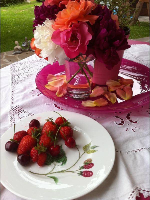 Farbenfrohe Blumen und frische Beeren auf einem Tisch im Freien, mit Garten im Hintergrund.