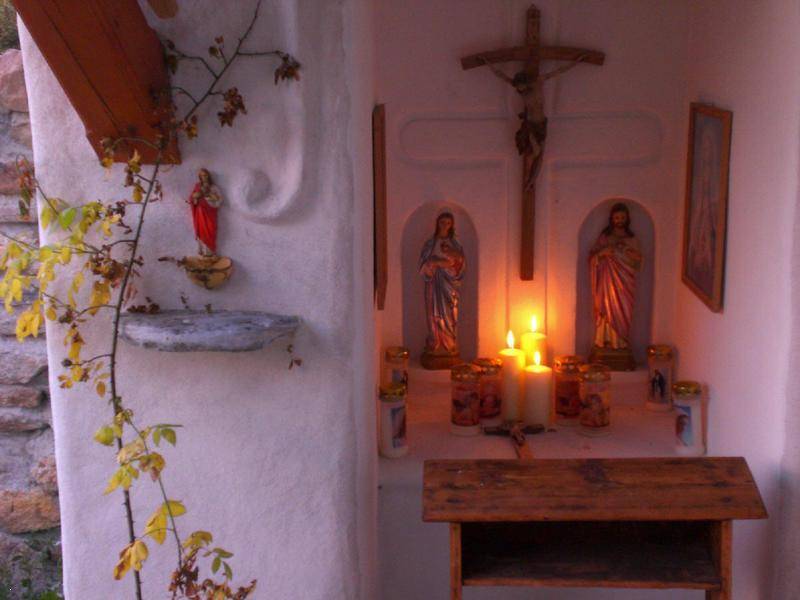 Beleuchtete Außennische mit religiösen Figuren, Kreuz und brennenden Kerzen auf einem Holztisch.