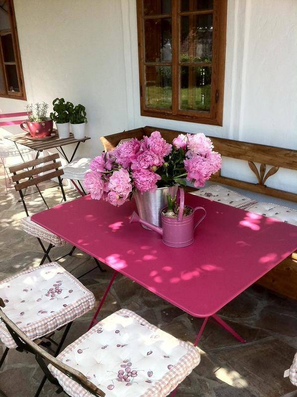 Sonnige Terrasse mit Holzsitzbank, rosa Tisch und einem Strauß frischer Pfingstrosen.