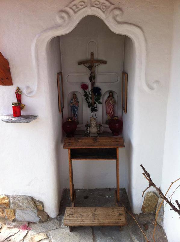 Detailansicht einer Außennische mit religiösen Figuren, Kreuz und einem kleinen Holztisch.