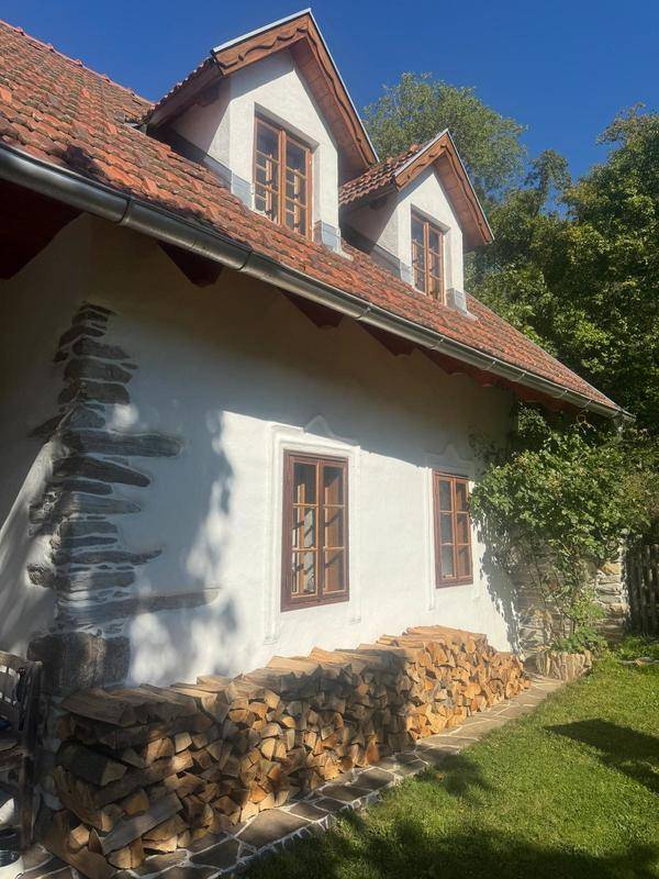 Traditionelles Landhaus mit weißer Fassade, roten Dachziegeln und gestapeltem Brennholz im Vorgarten.