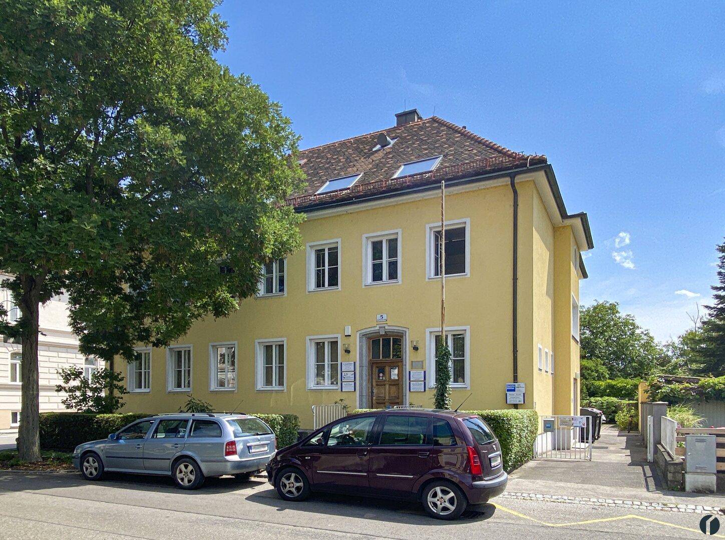 Ansicht des gelben Gebäudes von der Straße, umgeben von Bäumen und Hecken, mit Parkmöglichkeiten.