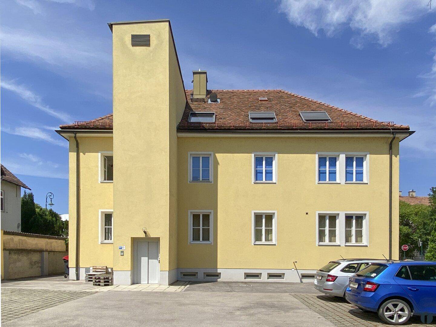 Gelbes Mehrfamilienhaus mit markantem Anbau und Parkplätzen vor dem Gebäude unter blauem Himmel.