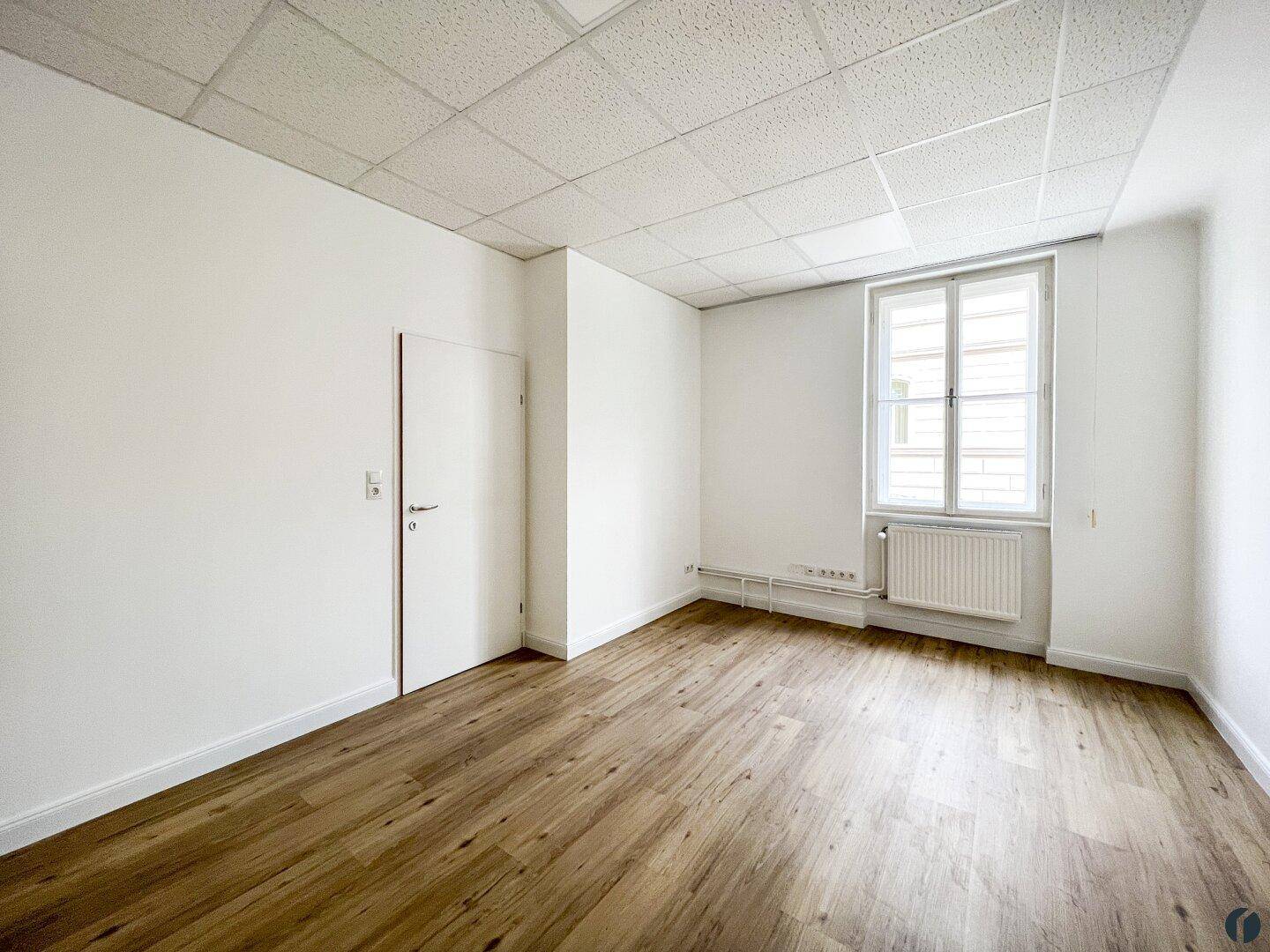 Leeres Büro mit Laminatboden, einem Fenster und weiß gestrichenen Wänden, bereit zur Einrichtung.