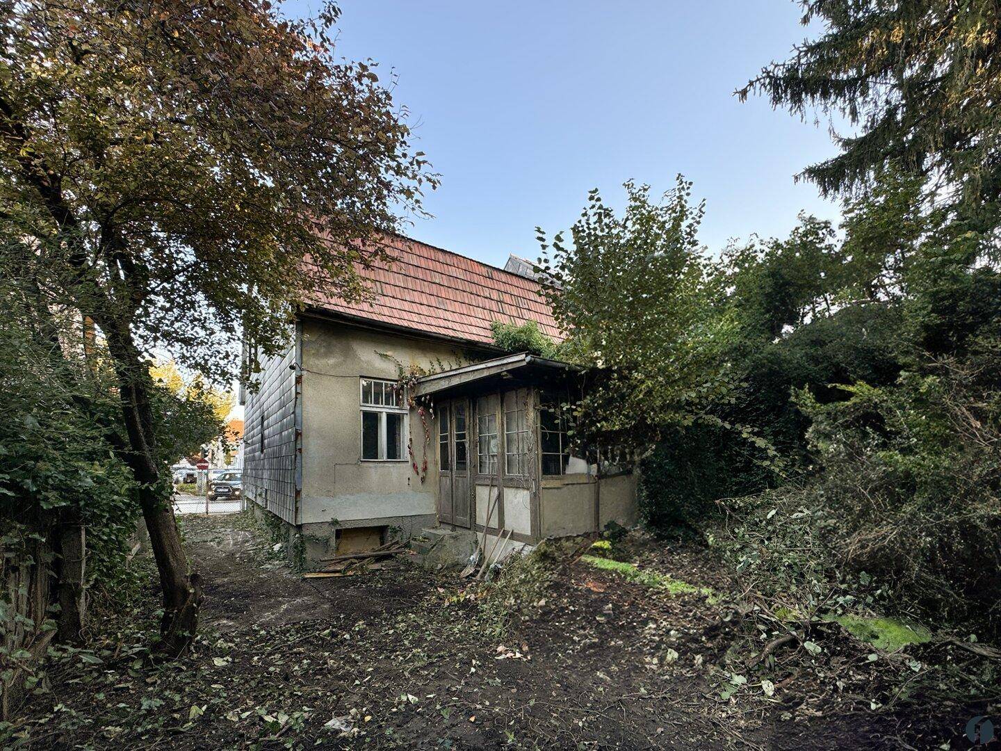 Altes Wohnhaus mit kleiner Veranda und verwildertem Garten, umgeben von Bäumen und Sträuchern.