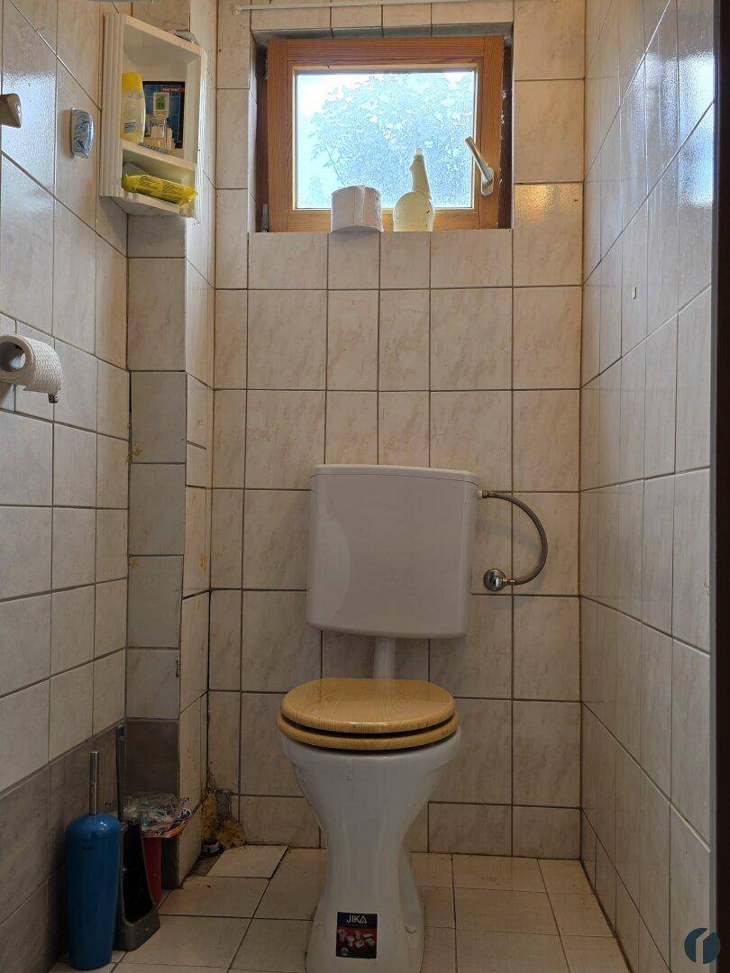 Kleiner Toilettenraum mit hellen Fliesen, einem Fenster und einem wandmontierten Spülkasten über der Toilette.