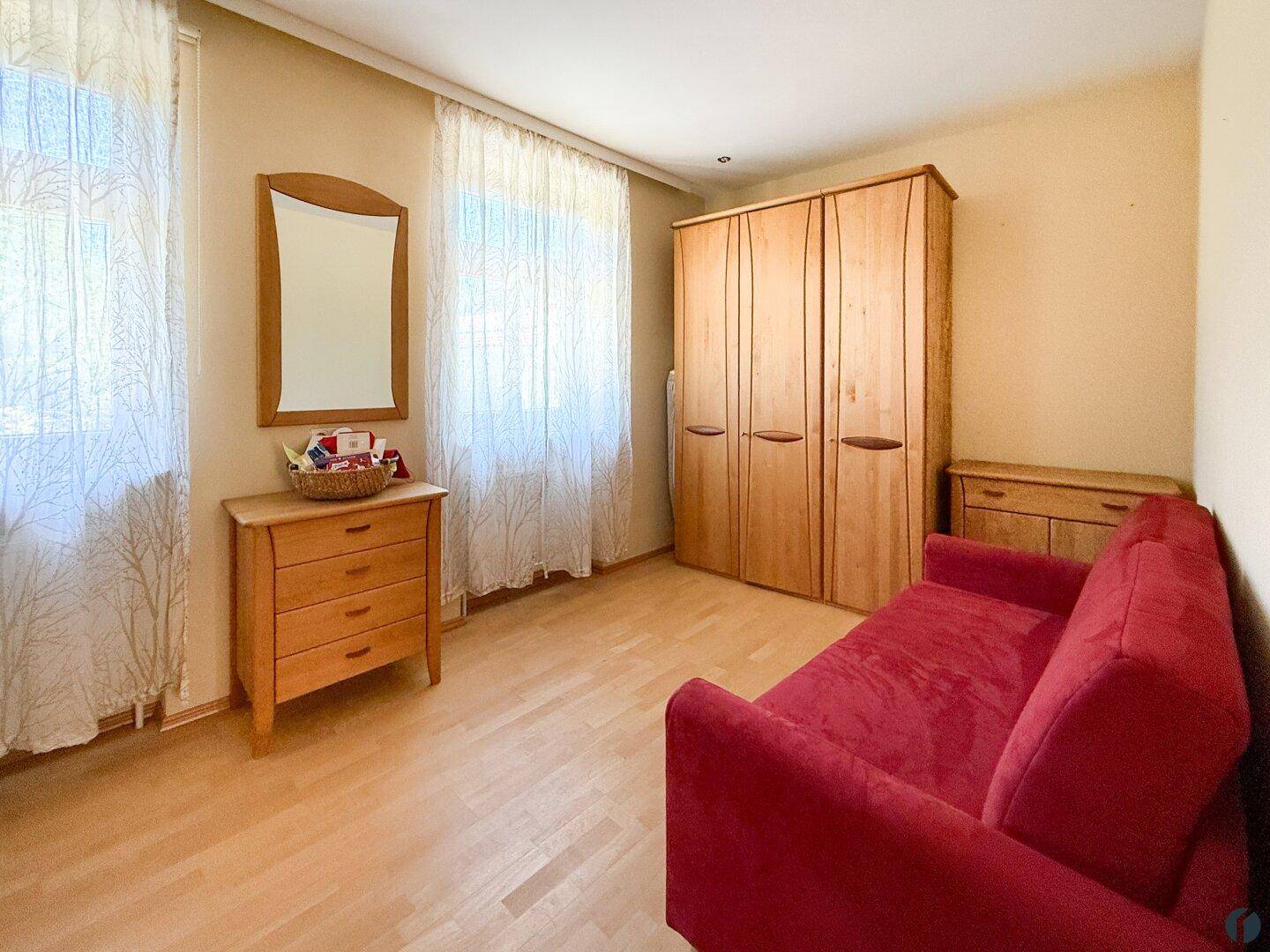Helles Zimmer mit Holzboden, rotem Schlafsofa und Holzschrank, ideal als Gästezimmer.