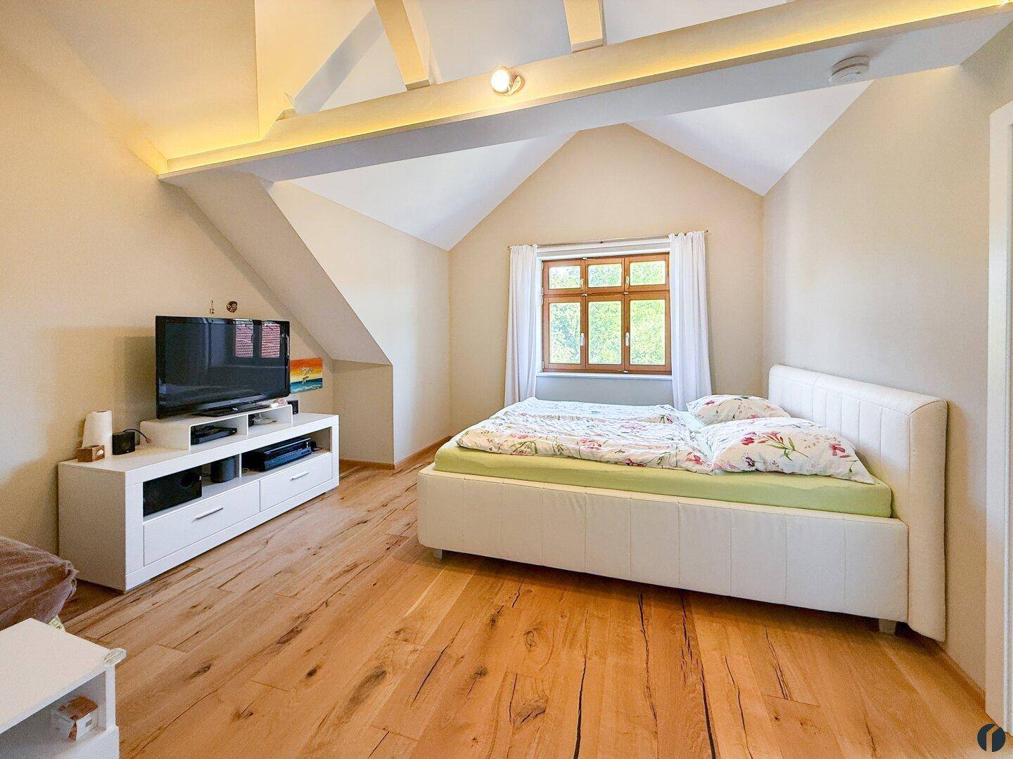 Helles Dachgeschoss-Schlafzimmer mit Doppelbett, TV-Möbel und sichtbaren Holzbalken.