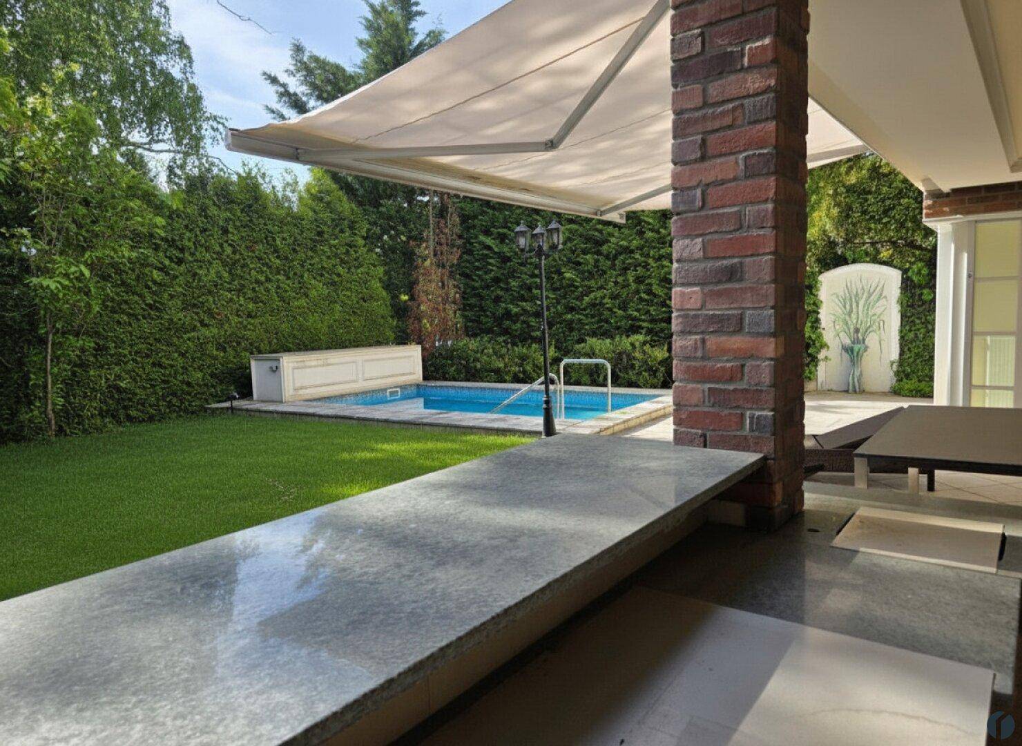 Gepflegter Gartenbereich mit Pool, Rasenfläche und überdachtem Sitzbereich unter einer Markise.