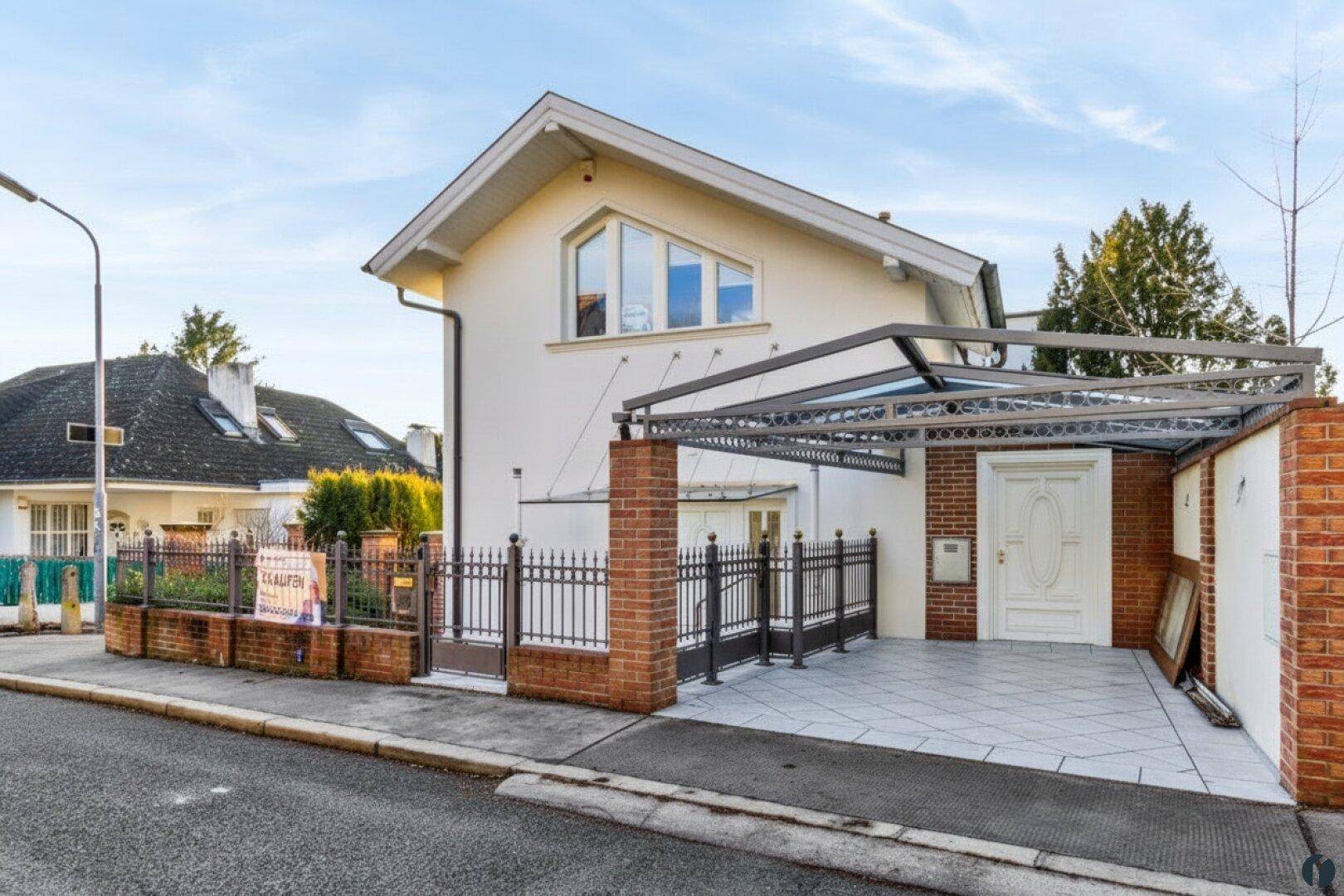 Einfamilienhaus mit Carport, gepflasterter Einfahrt und schmiedeeisernem Zaun an der Straße.