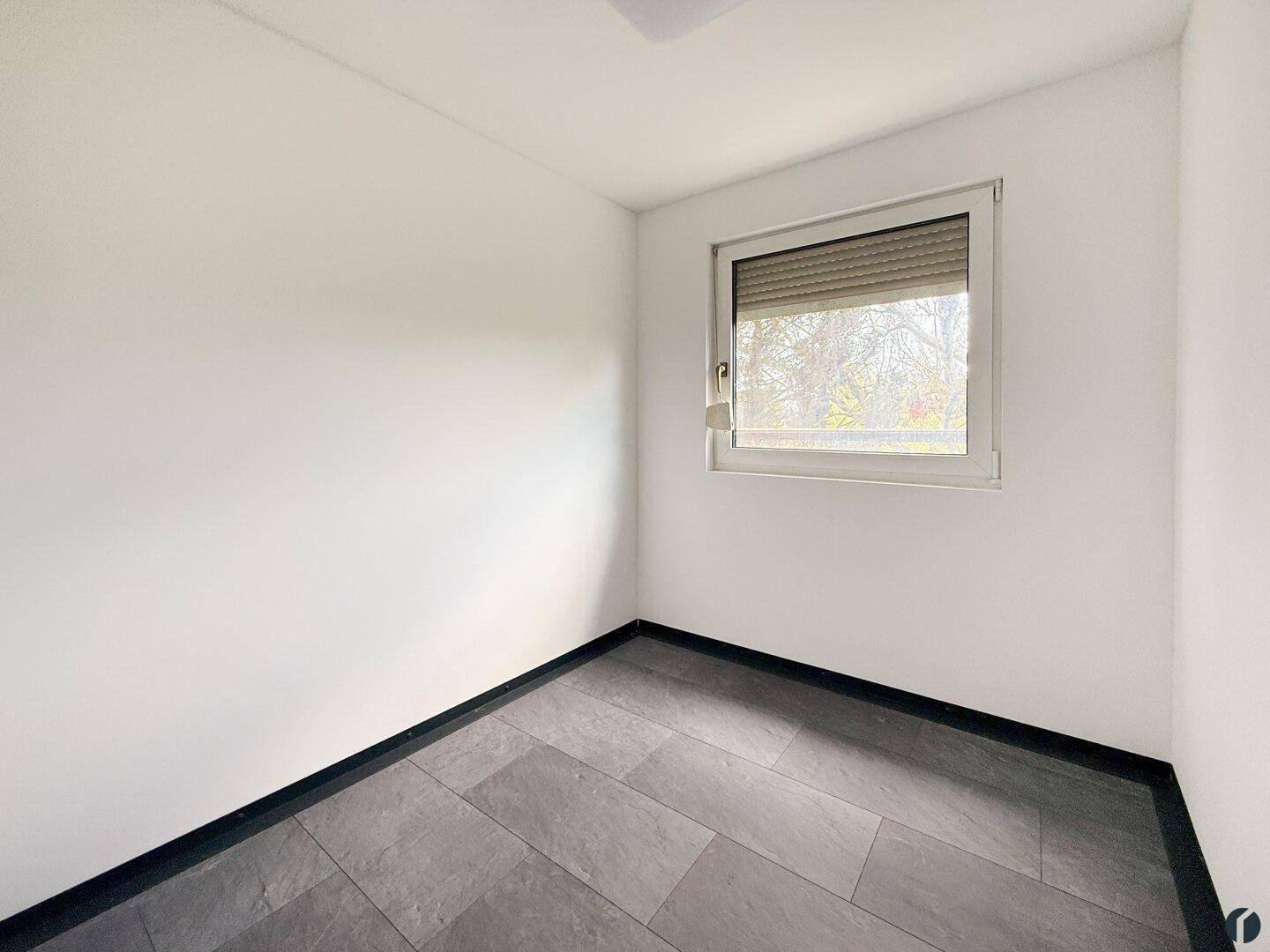 Kleines, helles Zimmer mit grauen Fliesen und einem Fenster mit Rollladen.