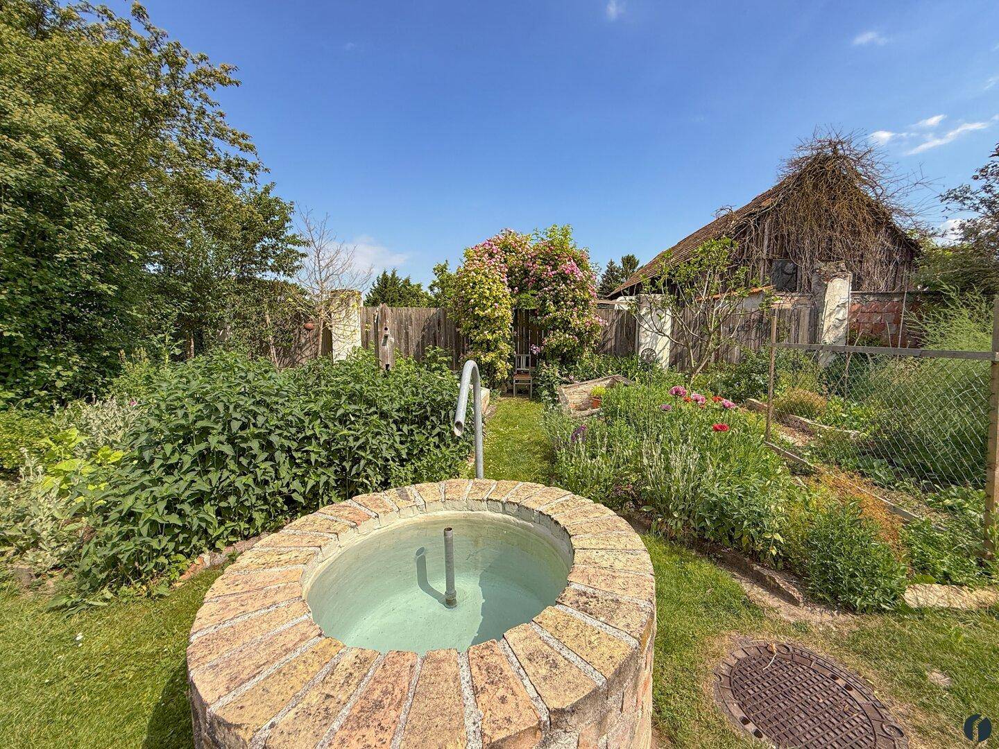 Gartenbereich mit einem gemauerten Brunnen, Blumenbeeten und üppiger Vegetation.
