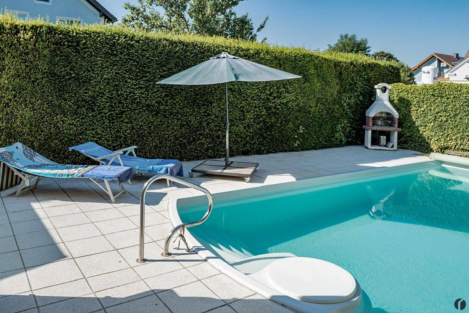 Außenbereich mit Swimmingpool, Liegestühlen, Grillplatz und hoher Hecke.