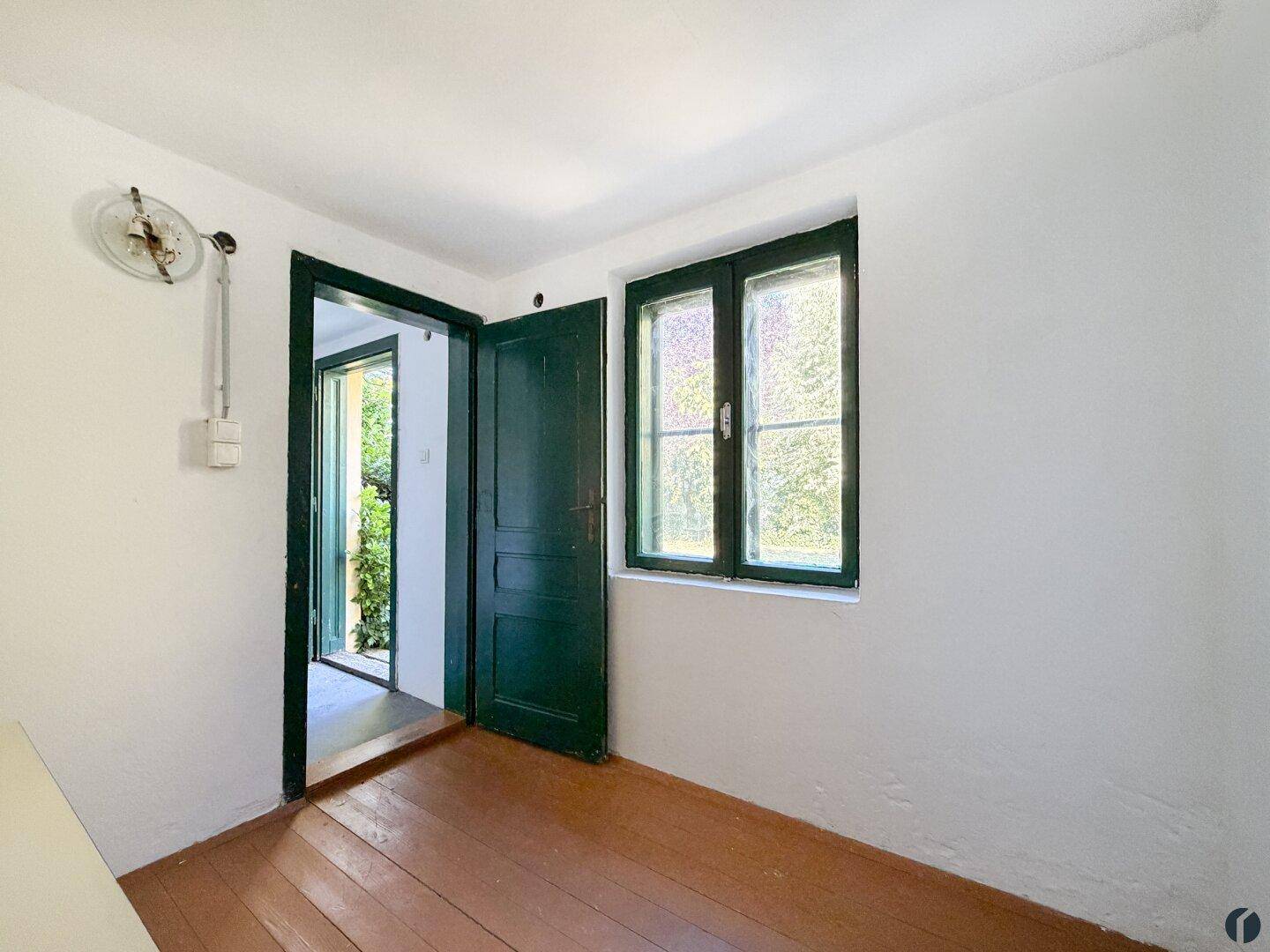 Kleiner Raum mit Holzboden und Fenster, das den Blick ins Grüne freigibt.
