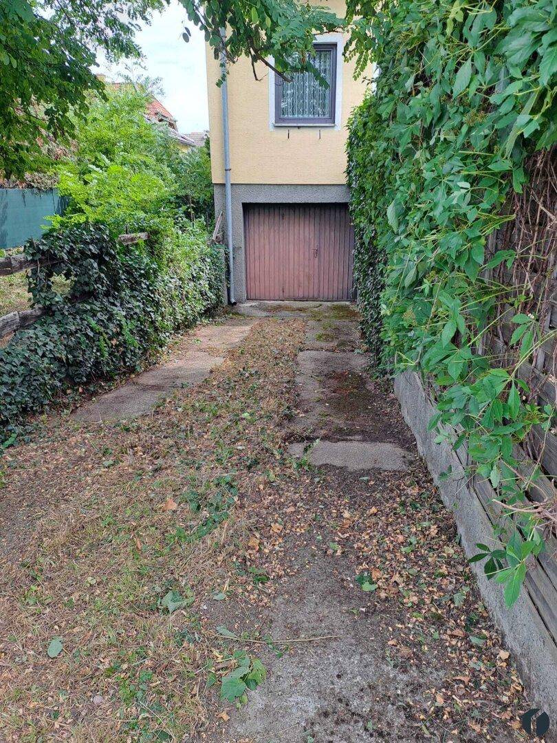 Einfahrt zu einer Garage mit Holztor, umgeben von grünen Hecken und Bäumen.