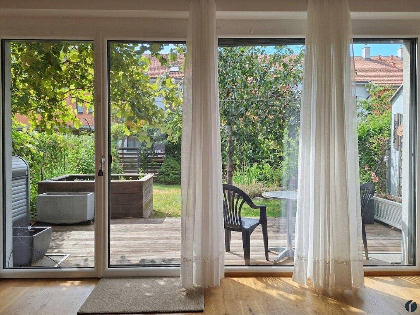 Blick aus dem Innenraum auf eine Holzterrasse und den grünen Garten.