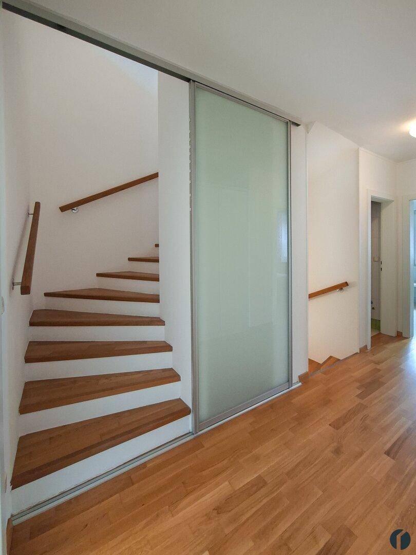 Heller Flur mit Holztreppe und moderner Glasschiebetür.
