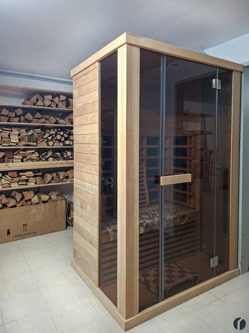 Moderne Sauna-Kabine aus Holz mit Glastür, umgeben von Holzscheiten im Kellerbereich.