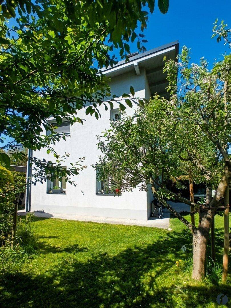 Helle Hausfassade umgeben von grünem Garten und blühenden Bäumen an einem sonnigen Tag.