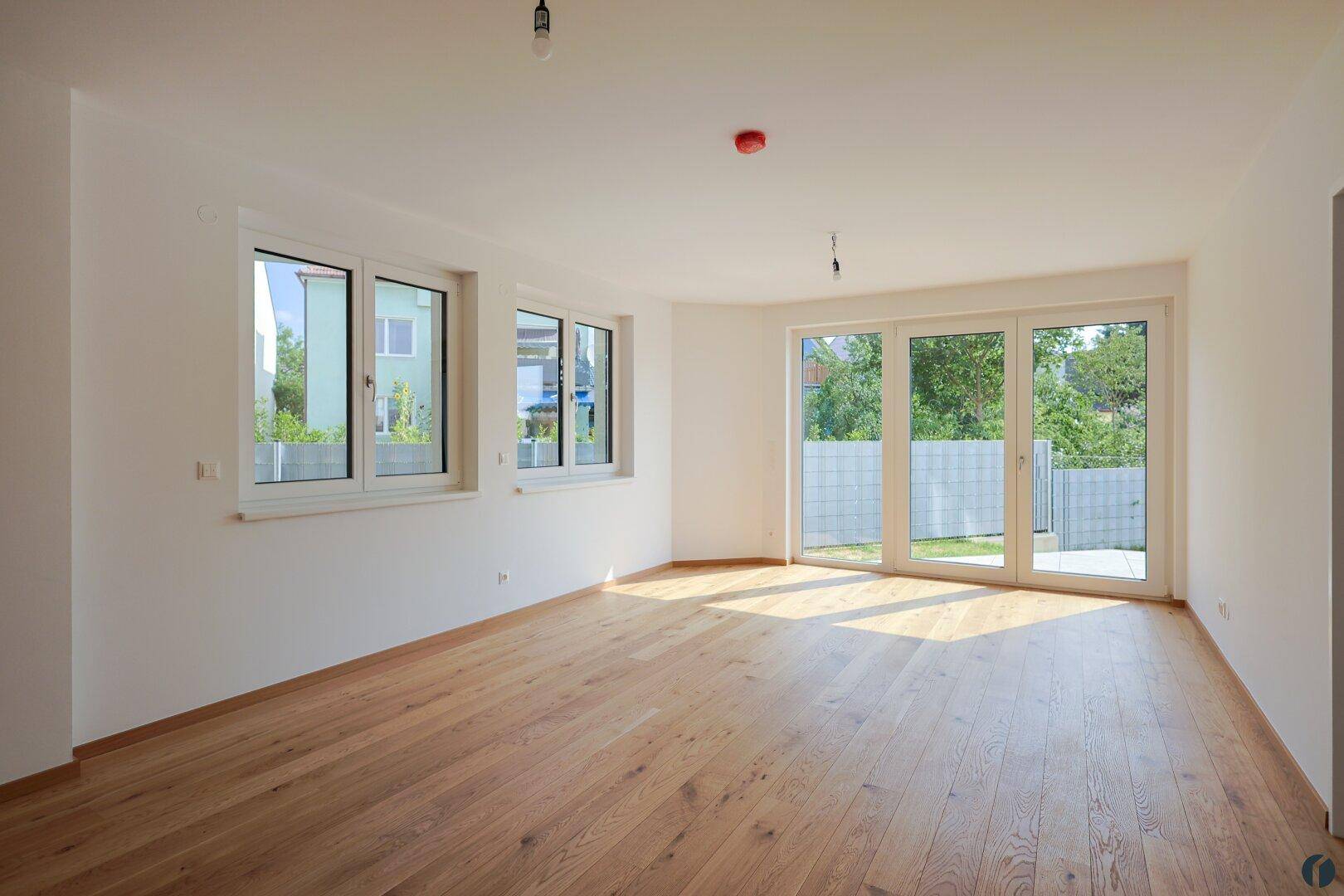 Geräumiges, leeres Wohnzimmer mit großen Fenstern und Zugang zum Garten, ausgestattet mit Holzboden.