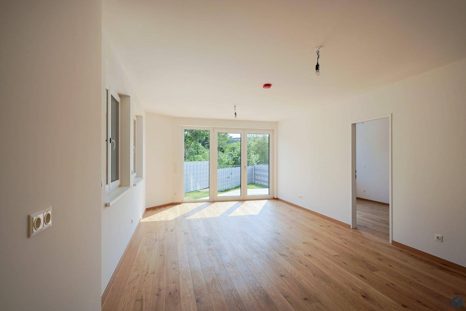 Helles, unmöbliertes Wohnzimmer mit Holzboden und großen Fenstern, die viel Tageslicht hereinlassen.