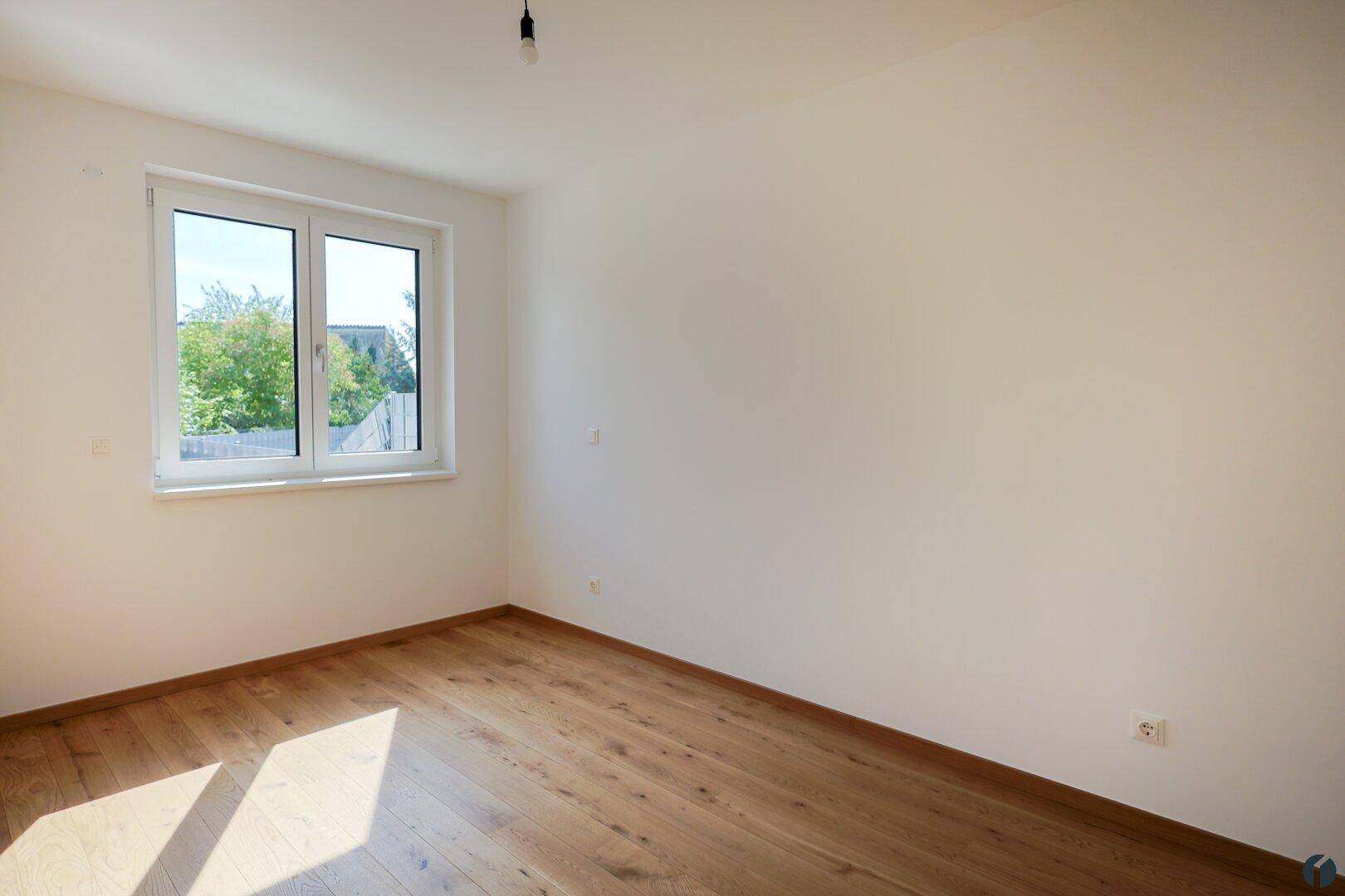 Leeres Schlafzimmer mit Holzboden und einem großen Fenster, das viel natürliches Licht spendet.