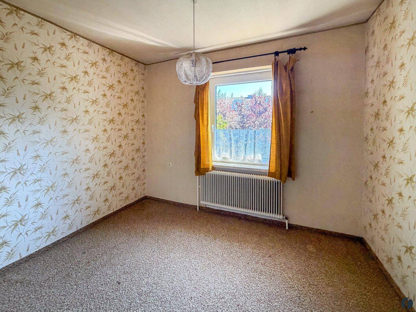 Schlafzimmer mit gemusterter Tapete, Teppichboden und einem Fenster mit Blick ins Grüne.
