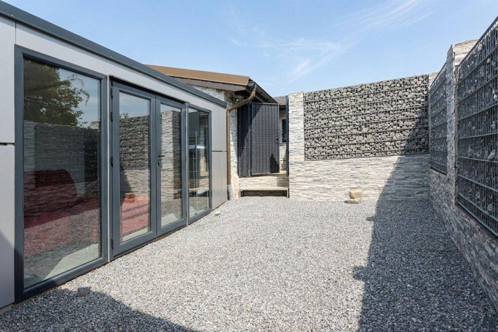 Außenbereich mit Kiesboden und modernen Glasfalttüren, umgeben von Gabionenwänden.