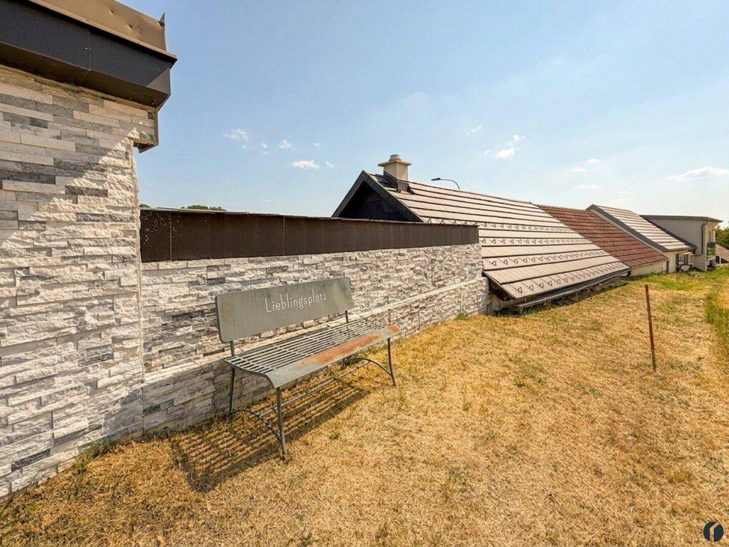 Außenbereich mit Steinmauer, einer Bank und Blick auf die Dächer der Nachbargebäude.
