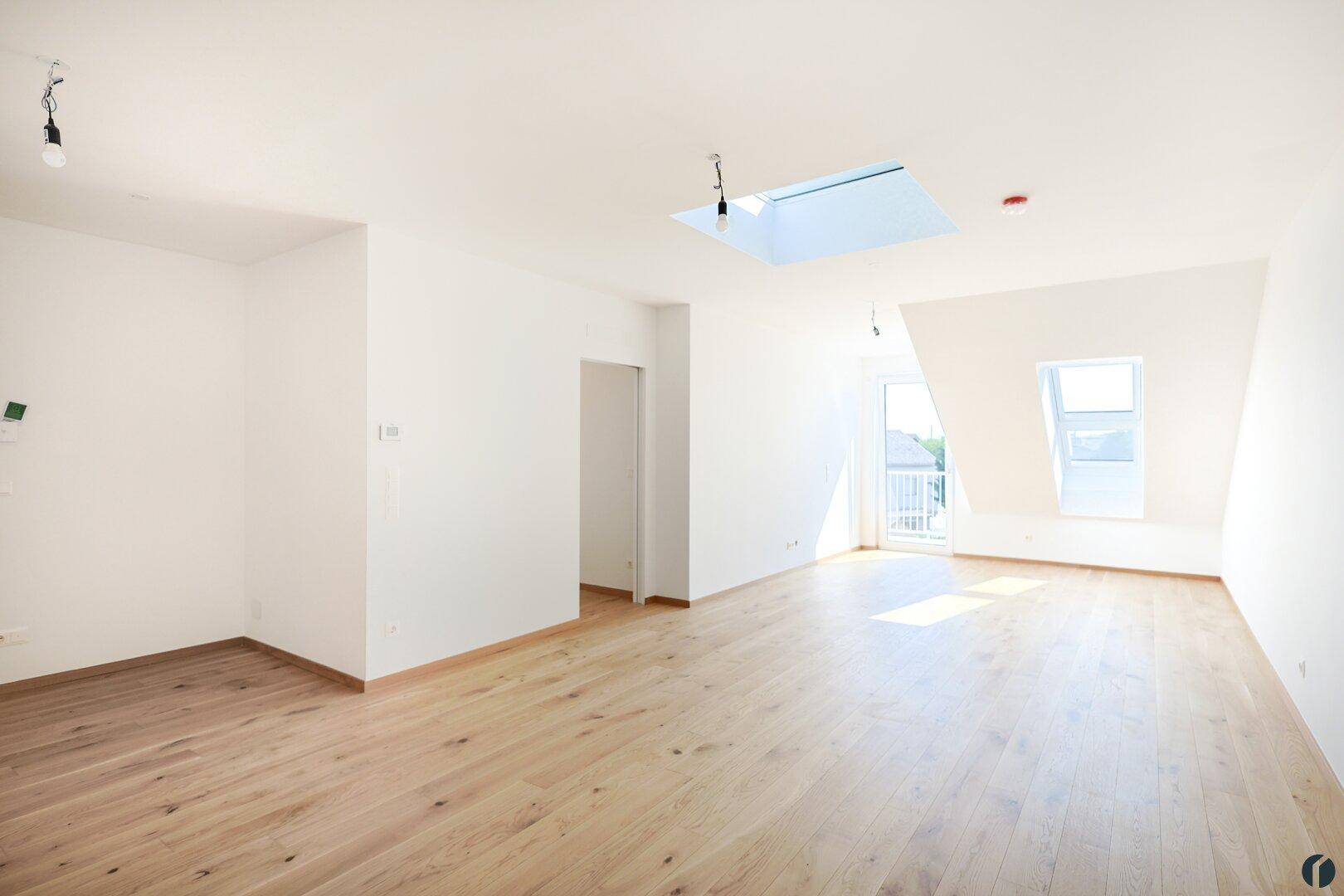 Heller, leerer Wohnbereich mit Holzboden, Dachfenster und Zugang zum Balkon.