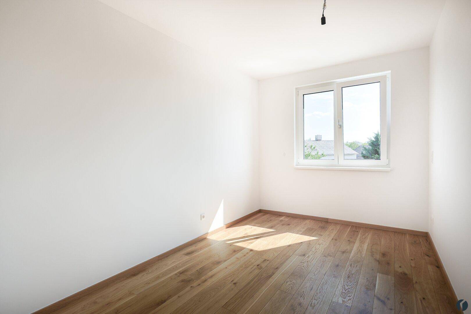 Helles, leeres Zimmer mit Holzboden und großem Fenster, ideal als Schlaf- oder Arbeitszimmer.