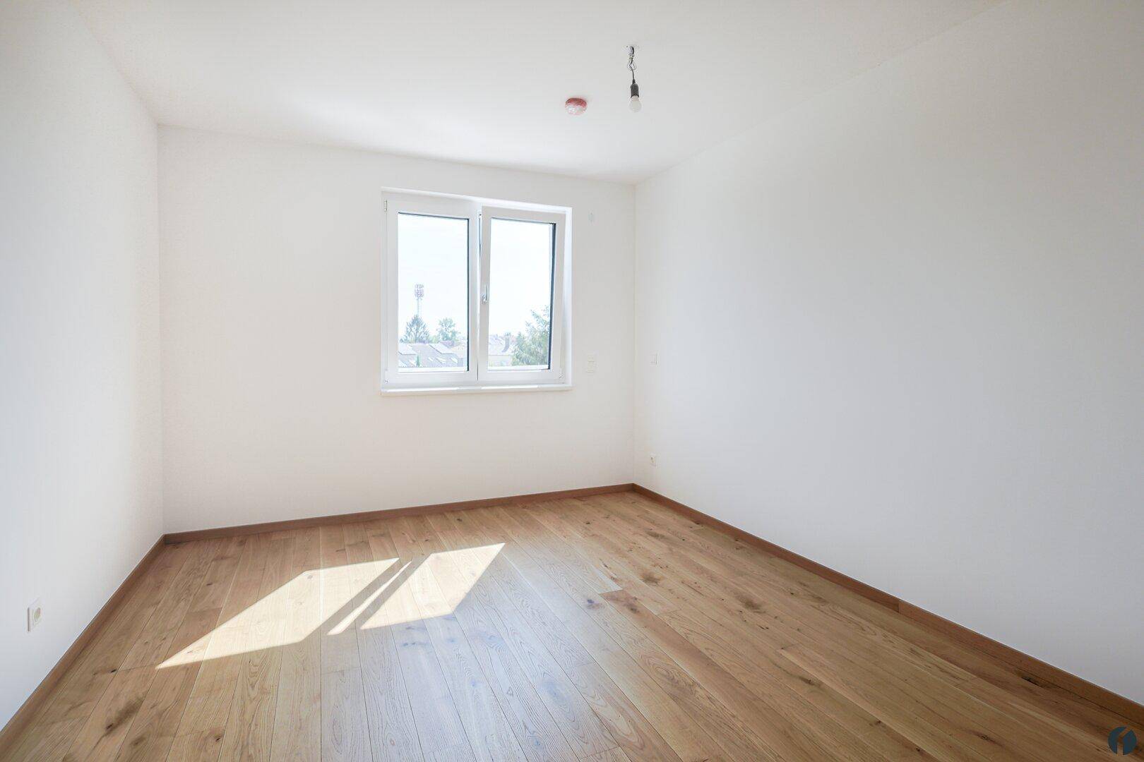 Geräumiges, leeres Zimmer mit Holzboden und viel Tageslicht durch das große Fenster.