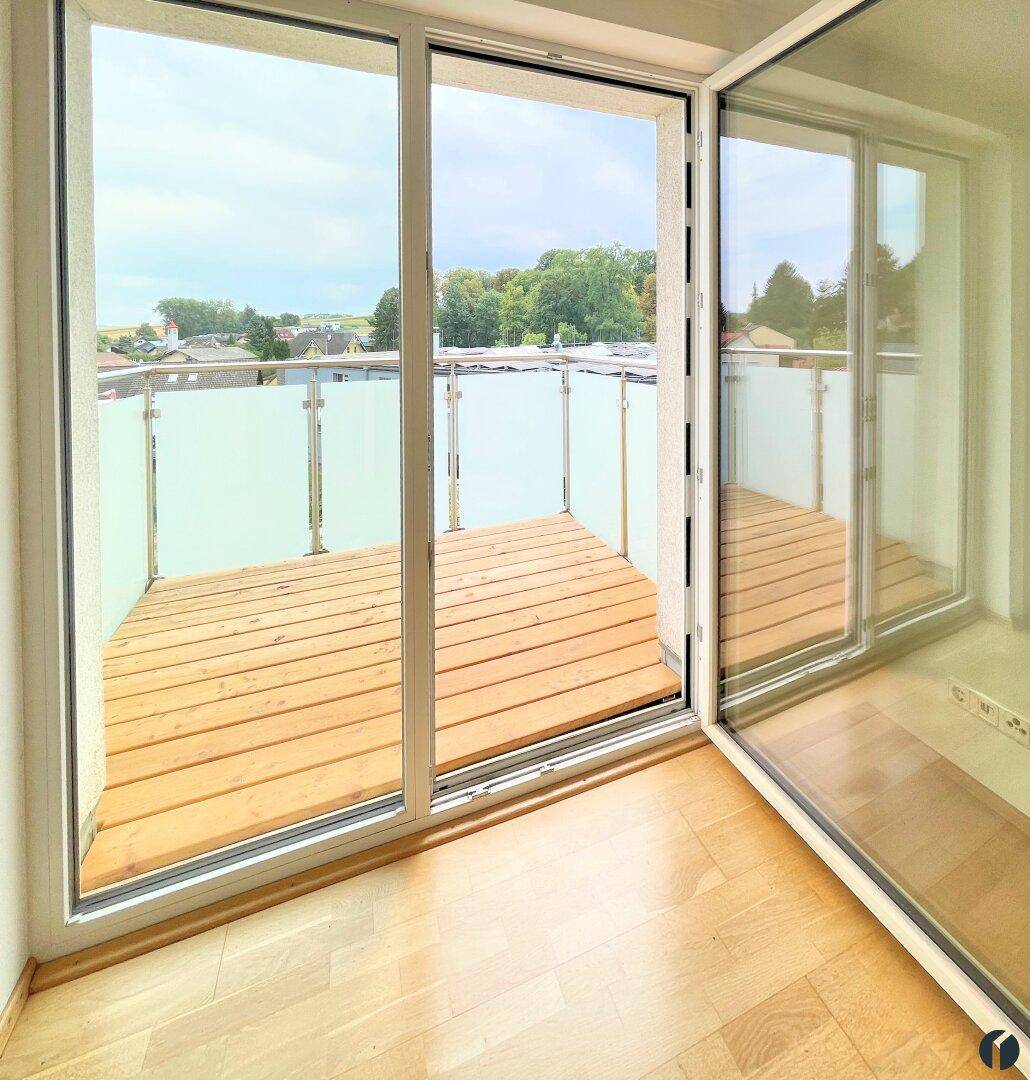 Geräumiger Balkon mit Holzboden, Glasgeländer und weitem Blick ins Grüne.