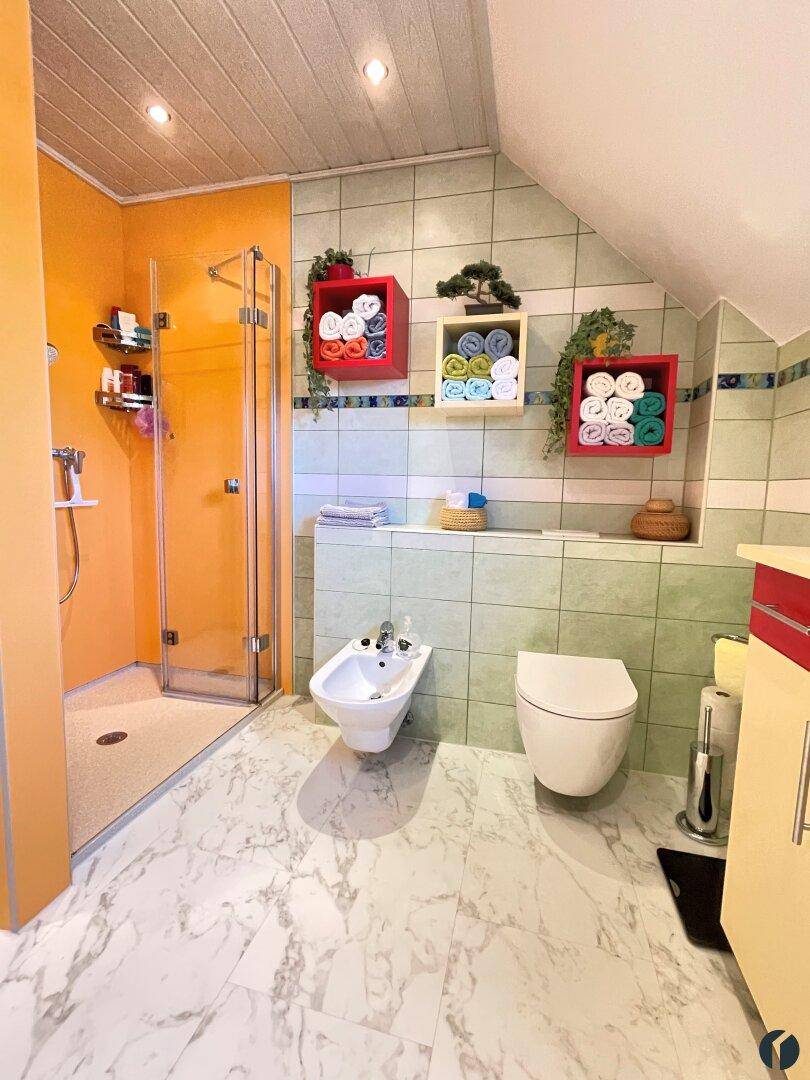 Badezimmer mit begehbarer Dusche, Bidet, WC und farbigen Fliesen, unter einer Dachschräge.