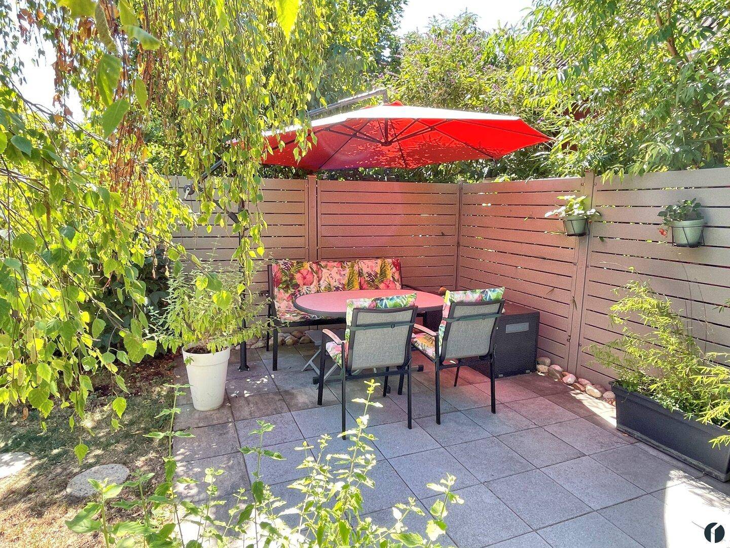 Gemütlicher Sitzbereich im Garten mit Pflastersteinen, Holzzaun und rotem Sonnenschirm.
