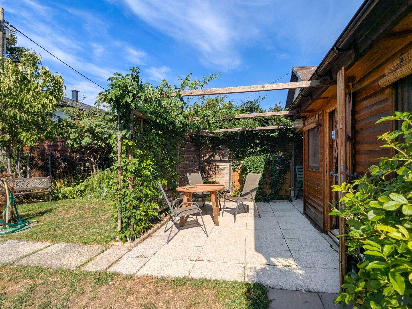 Sonniger Terrassenbereich mit Gartenmöbeln und einer begrünten Pergola neben dem Holzhaus.