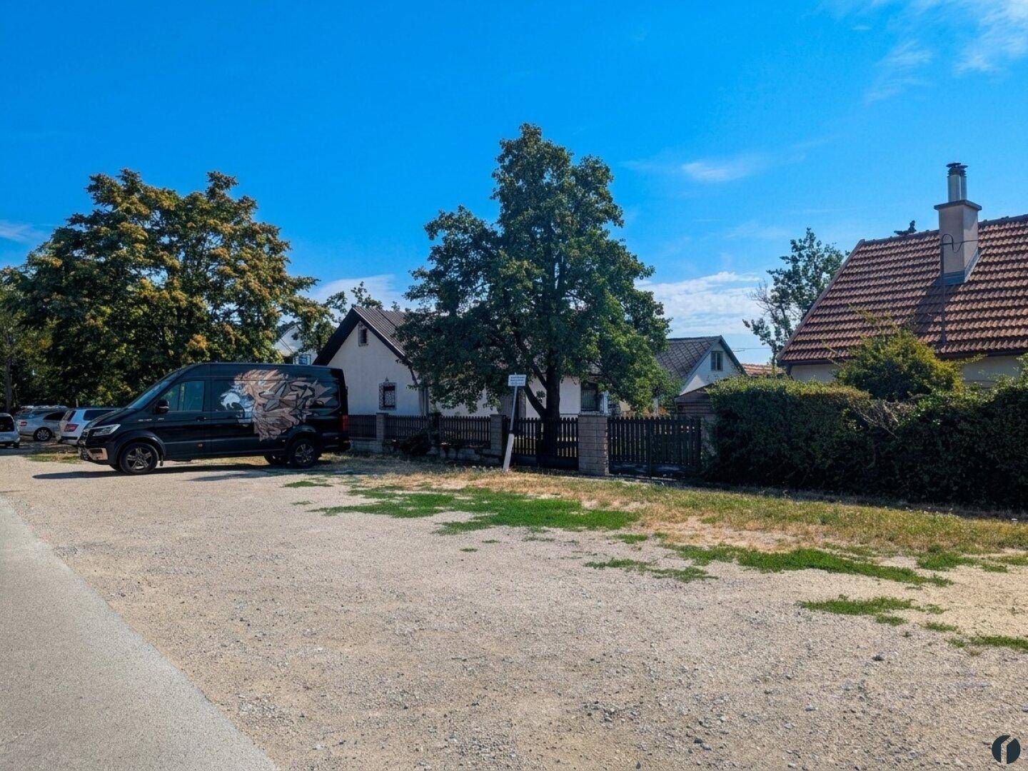 Außenansicht mit Parkplatz und umliegenden Häusern und Bäumen unter blauem Himmel.