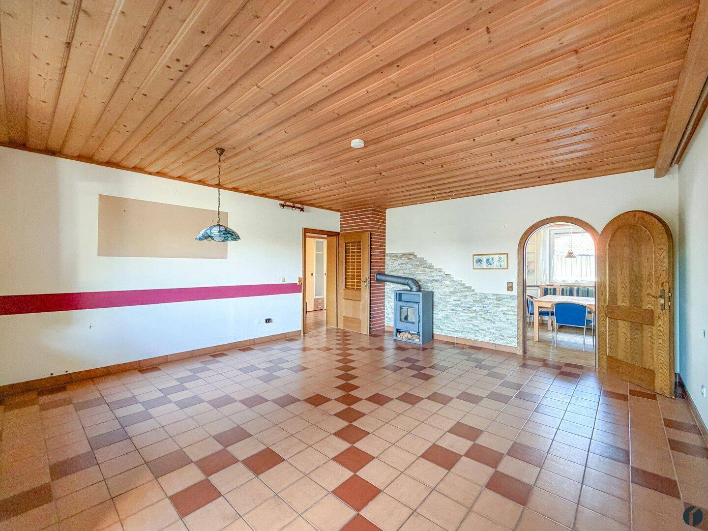 Helles Wohnzimmer mit Kaminofen, Fliesenboden und Holzbalkendecke.