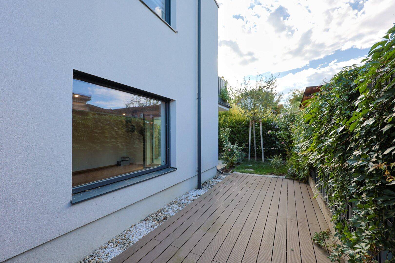 Seitlicher Außenbereich mit Holzdeck, Kiesstreifen und grüner Hecke entlang der Hauswand.