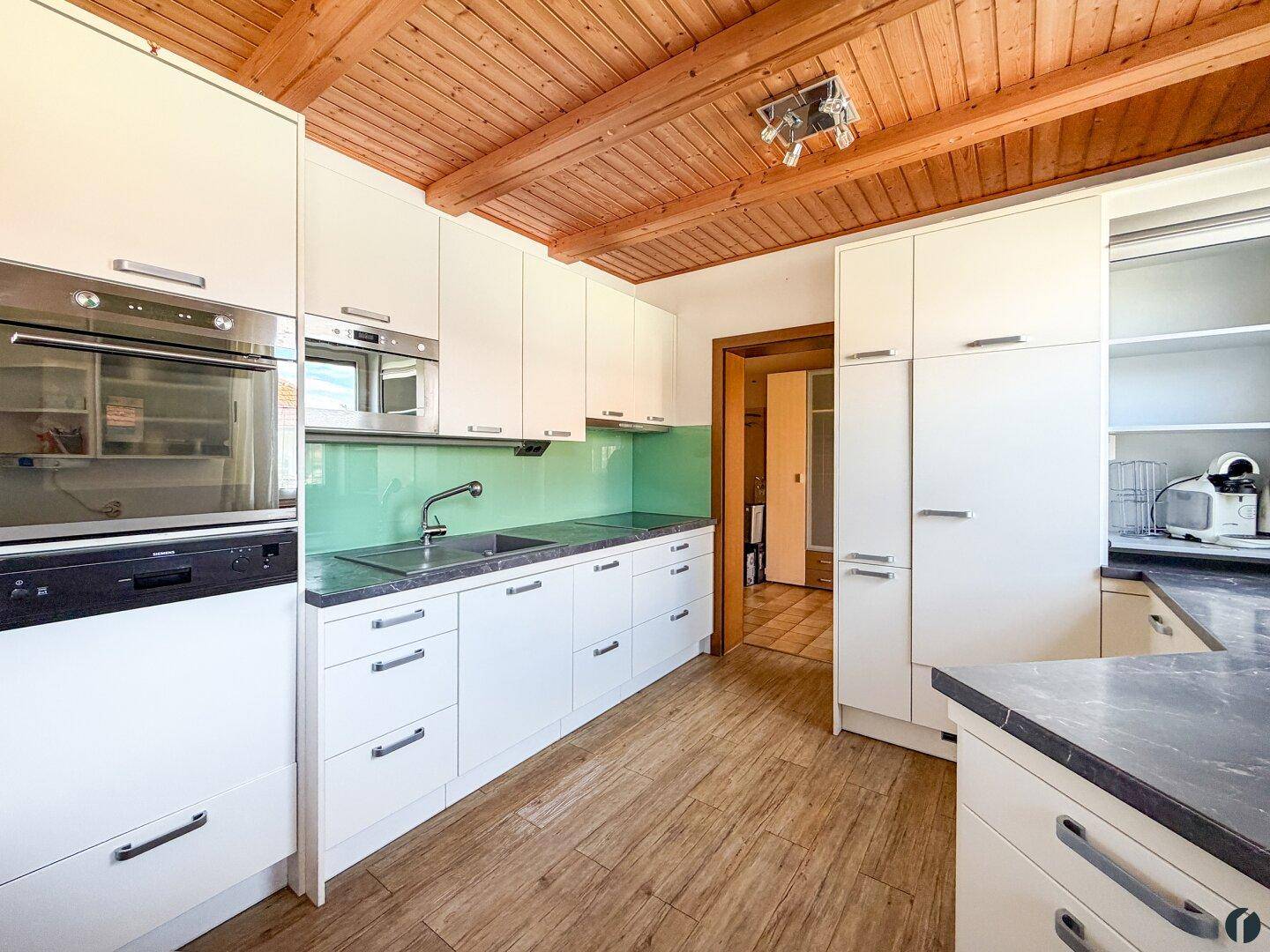 Moderne Einbauküche mit weißen Schränken, dunkler Arbeitsplatte und Holzbalkendecke.