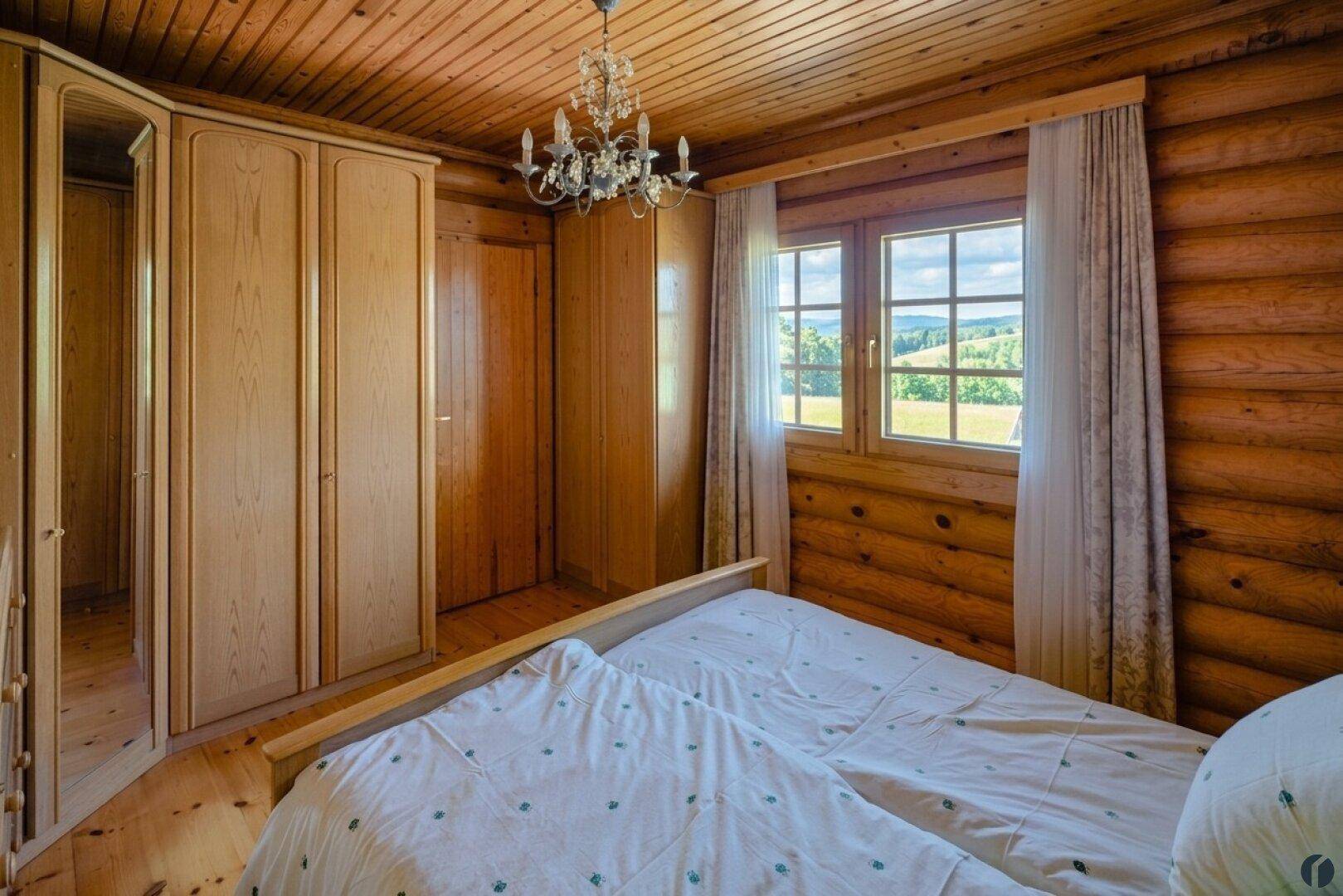 Schlafzimmer im Blockhaus mit Doppelbett, Holzschränken und Blick auf die grüne Landschaft.