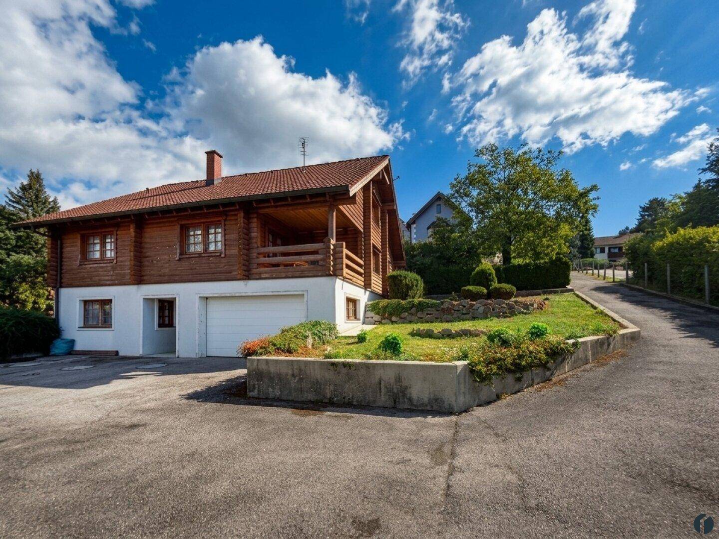 Blockhaus mit integrierter Garage und Satteldach, umgeben von gepflegtem Gartenbereich.