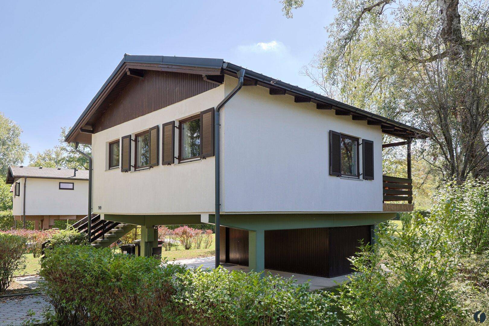 Seitliche Ansicht eines Stelzenhauses mit Fensterläden und einer Außentreppe in grüner Umgebung.