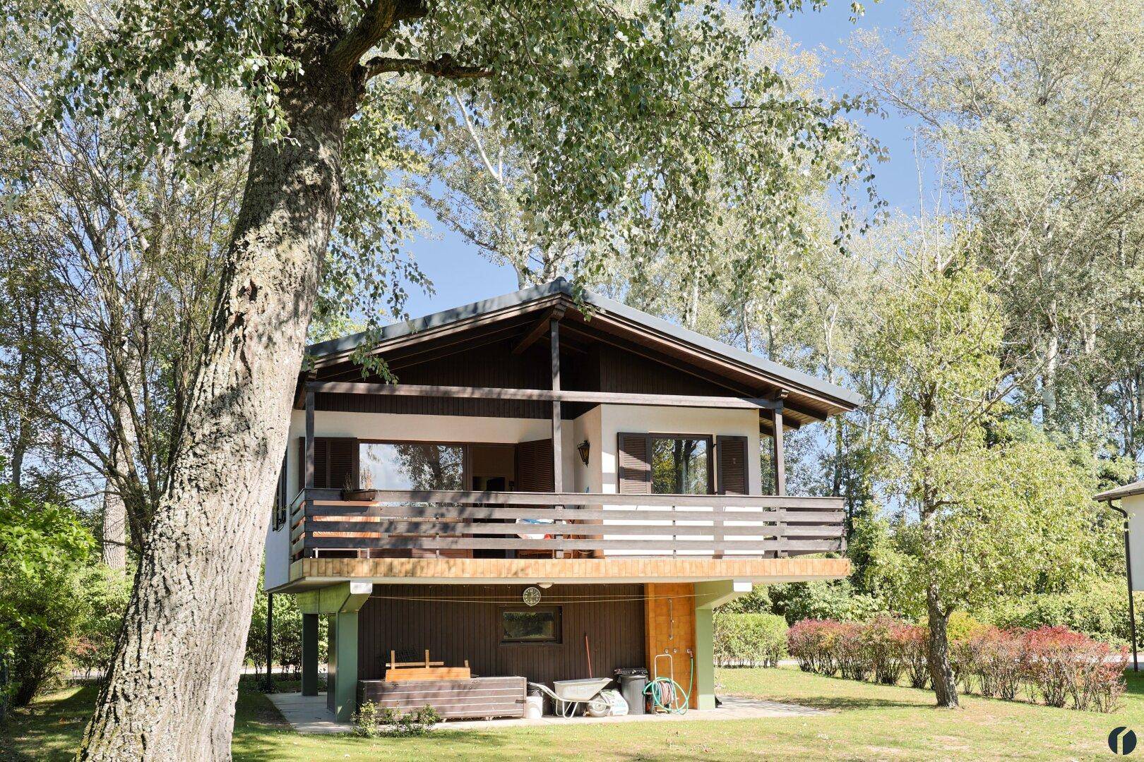 Charmantes Stelzenhaus mit Balkon, umgeben von hohen Bäumen und einem offenen Untergeschoss.