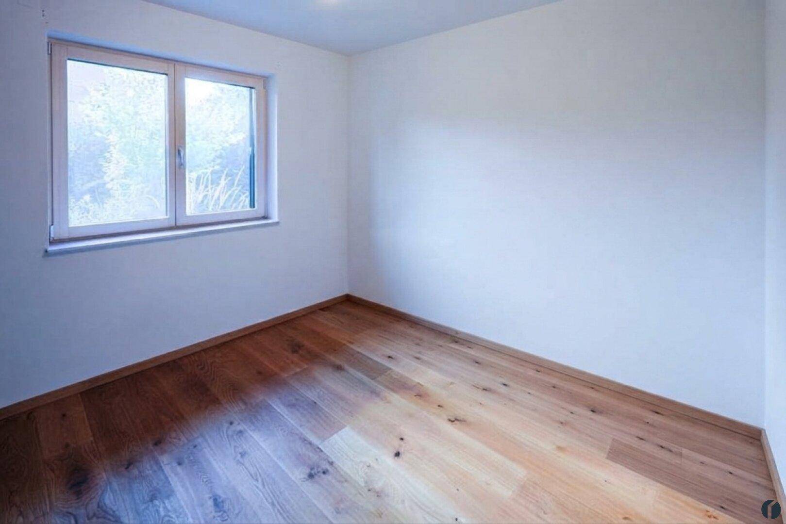 Lichtdurchflutetes Zimmer mit hellem Holzparkett und Blick ins Grüne durch das Fenster.