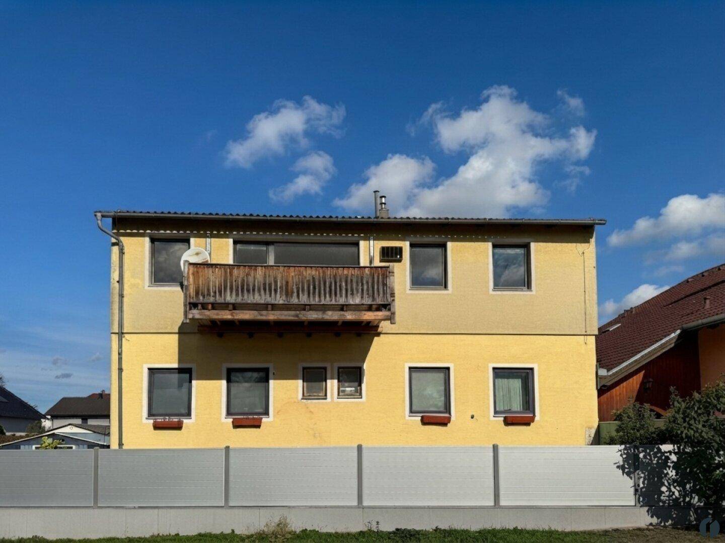 Gelbes Gebäude mit einem Holzbalkon und mehreren Fenstern, umgeben von einem Zaun.