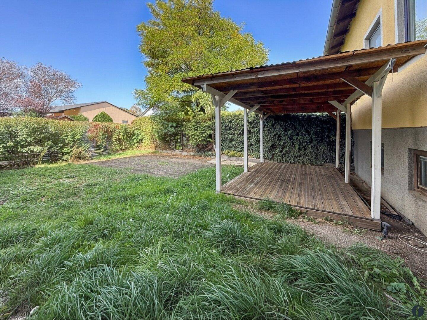 Gartenbereich mit einer überdachten Holzterrasse, Rasenfläche und grünen Hecken.